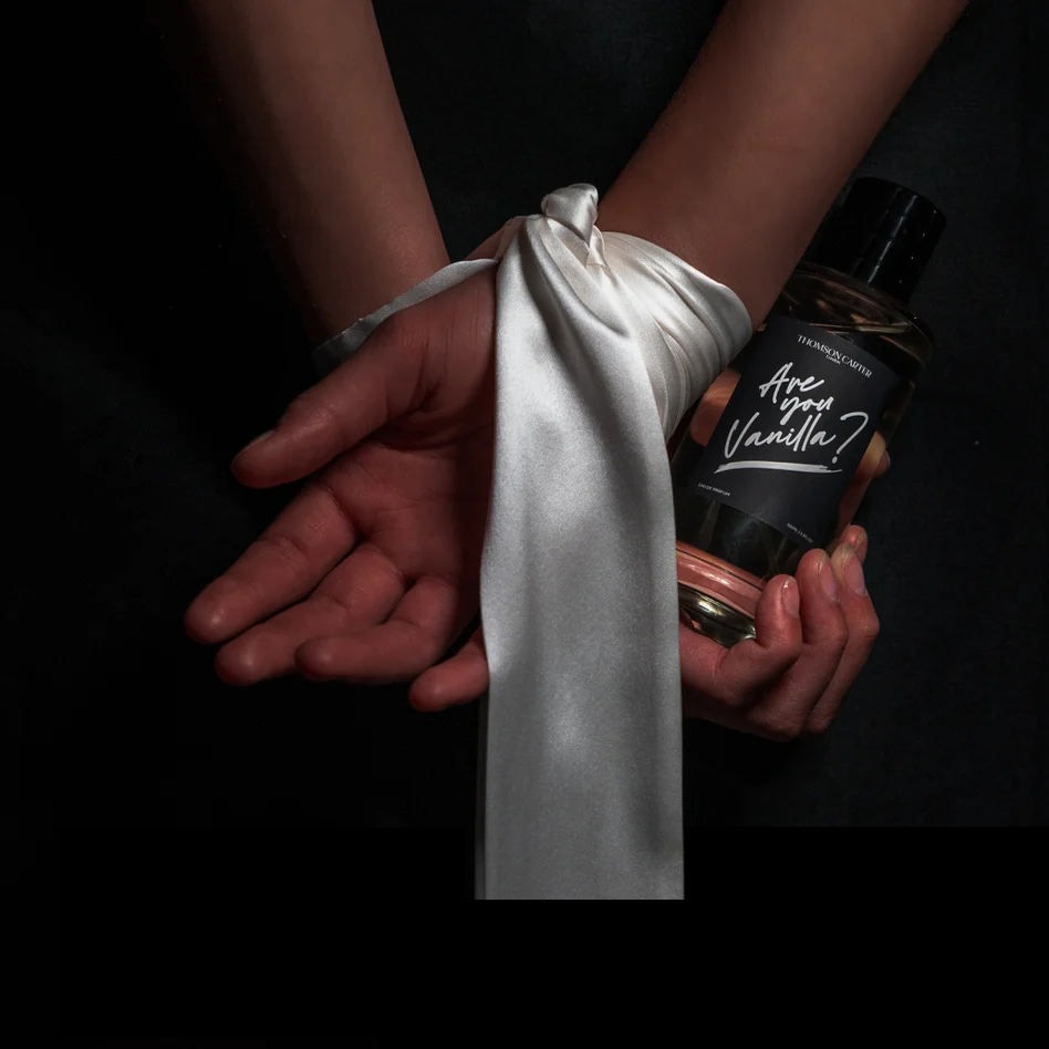 A person holds a bottle of Aphrodite Noir Eau de Parfum next to their wrist, which is wrapped in a shiny white satin ribbon. The background is solid black.