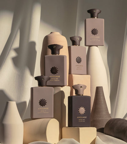 Six Arrouage body wash bottles in muted taupe, beige, and charcoal gray tones, arranged on geometric pedestals with draped fabric and soft shadows.