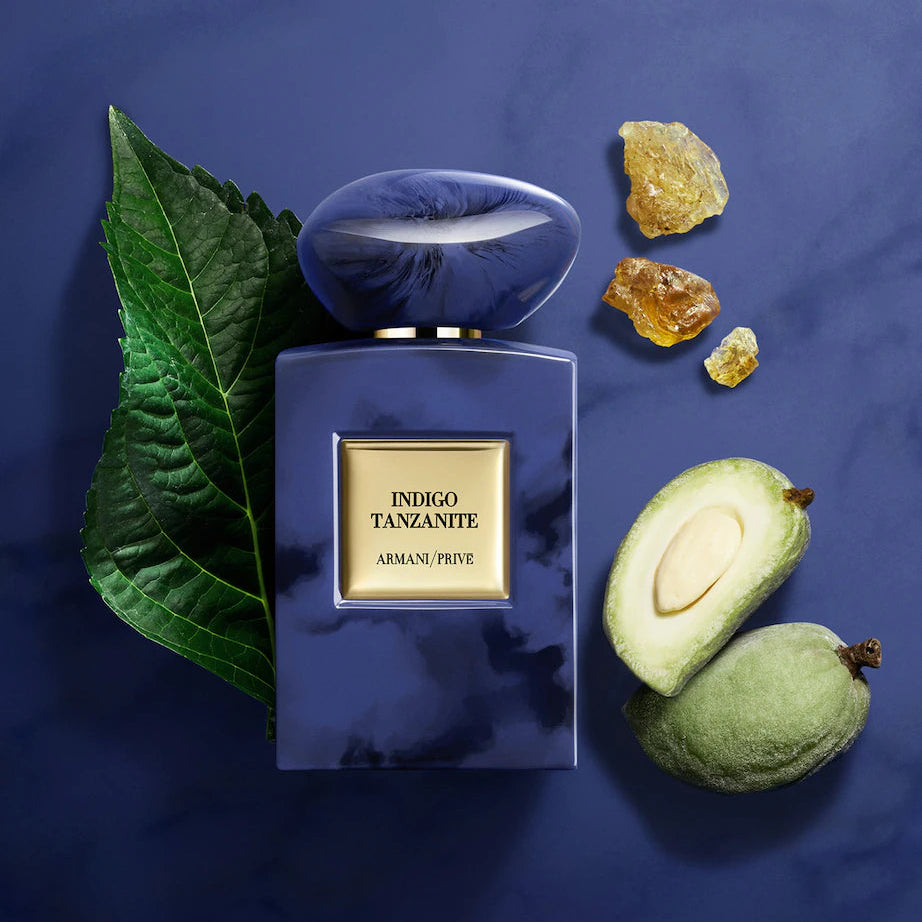 Armani Privé Indigo Tanzante perfume bottle with a blue marbled finish and gold label, styled with a green leaf, avocado halves, and amber crystals on a dark blue background.