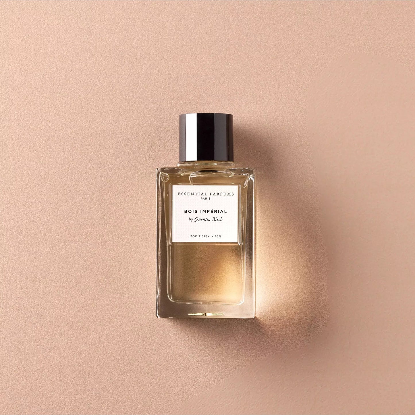 A rectangular glass perfume bottle with a black cap and a white label reading 'ESSENTIALS PARFUMER BOIS IMPERIAL', set against a warm beige textured background.
