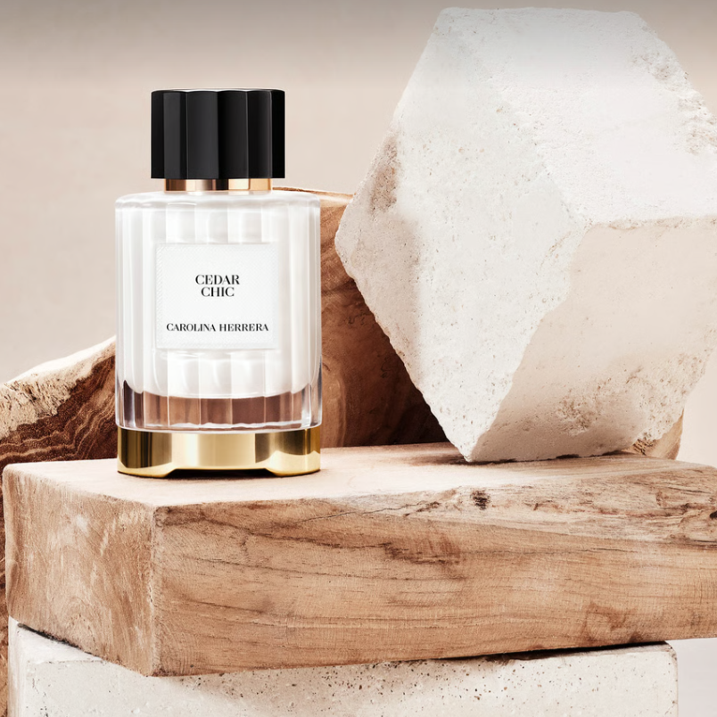A perfume bottle labeled 'CEDAR CHIC' by Carolina Herrera, featuring a white striped glass body, a black cap, and a gold base, styled on a rustic wooden block with textured stone props against a warm beige background.