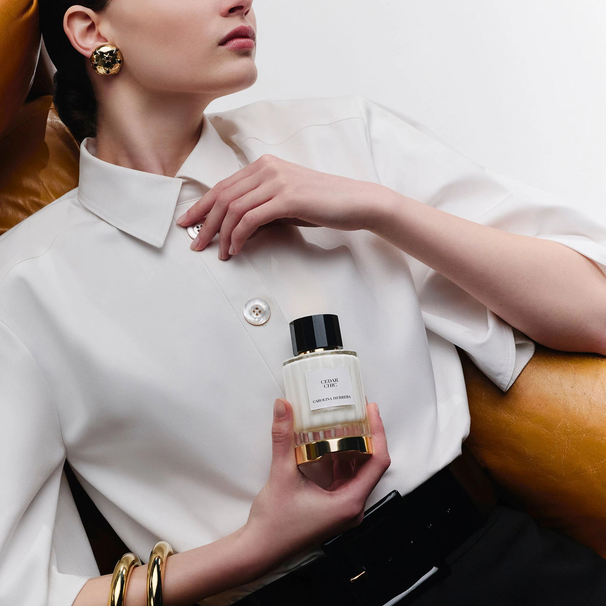A model in a white short-sleeve collared shirt holds a bottle of Tom Ford Tobacco House perfume, posed on a brown leather couch.