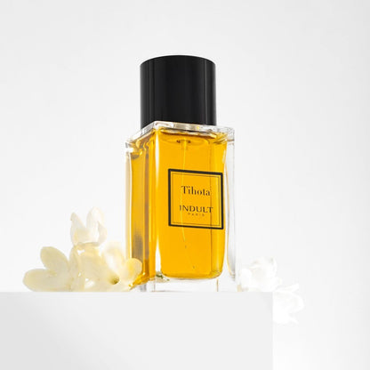 A clear rectangular glass bottle of Tihota Indult perfume with golden-yellow liquid, a black cap, and a black label, styled on a white pedestal with white flowers against a white background.