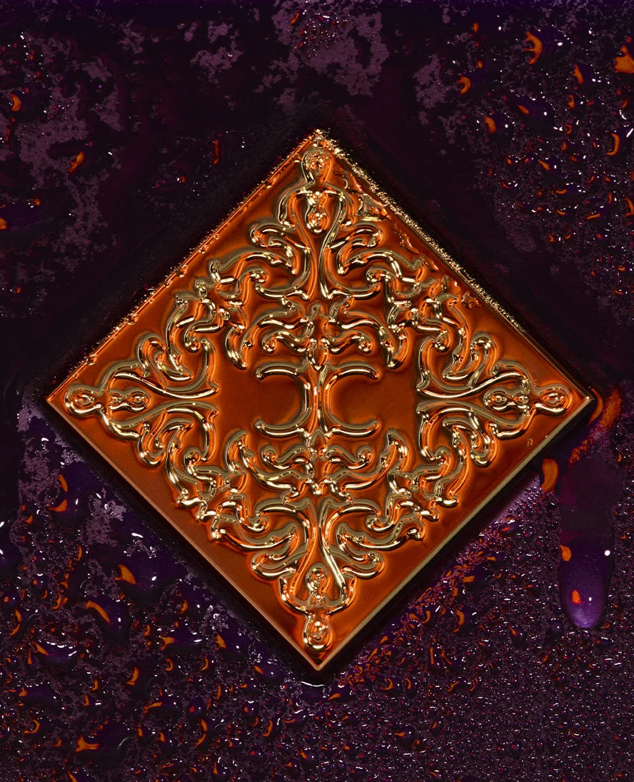 A square decorative tile with an ornate embossed floral pattern, shown at an angle on a dark, wet, textured surface.