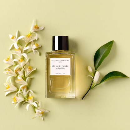 A rectangular glass perfume bottle with a black cap and a white label reading 'ESSENTIAL PERFUMES NEROLI BOTANICA' is arranged on a pale yellow-green background with white neroli flowers and green leaves around it.