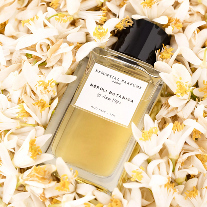 A rectangular glass perfume bottle filled with pale yellow liquid and topped with a black cap, surrounded by white neroli flowers. The front label reads 'Essential Parfums London' and 'Neroli Botanica by Anne-Heigo'.