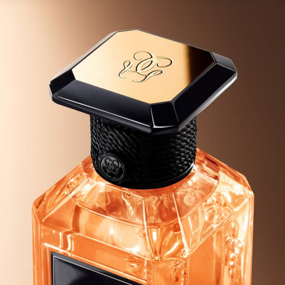A close-up of a luxury perfume bottle with a faceted amber-orange glass body, a textured black neck, and a geometric black cap featuring a gold top panel with an elegant monogram logo.