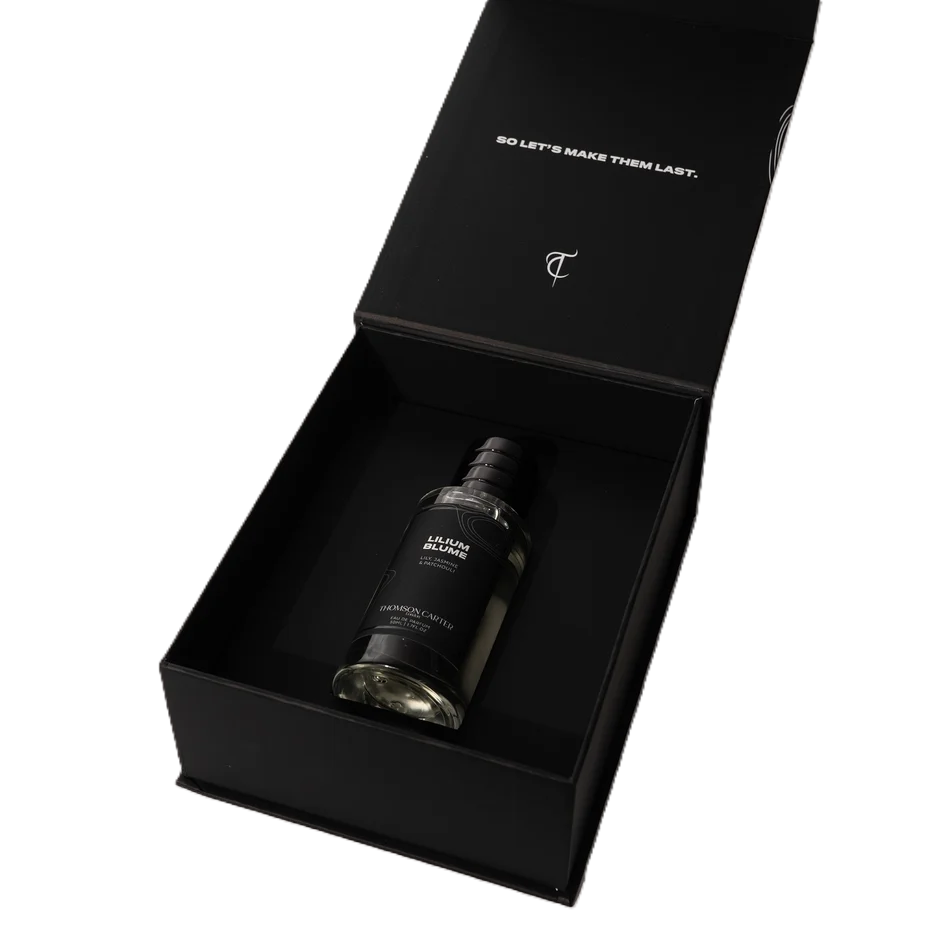 A black rectangular gift box with its lid open, containing a dark glass bottle labeled 'BURNING CANDLE'. The inside of the lid has white text reading 'SO LET'S MAKE THEM LAST.