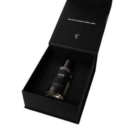 A black rectangular gift box with its lid open, containing a dark glass bottle labeled 'BURNING CANDLE'. The inside of the lid has white text reading 'SO LET'S MAKE THEM LAST.