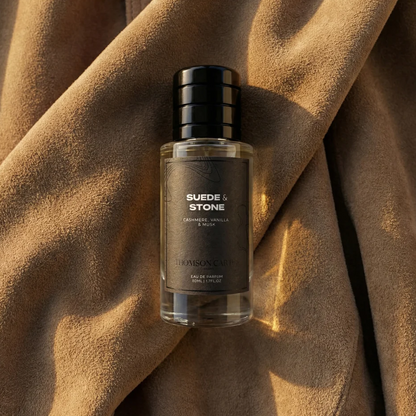 A bottle of Thomson carter Suede & Stone cologne with a black cap and dark gray label, resting on draped brown suede fabric.