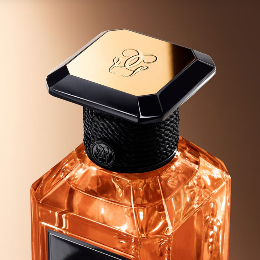A close-up of a faceted amber-orange perfume bottle with a black textured neck and a geometric gold cap featuring an engraved monogram logo.