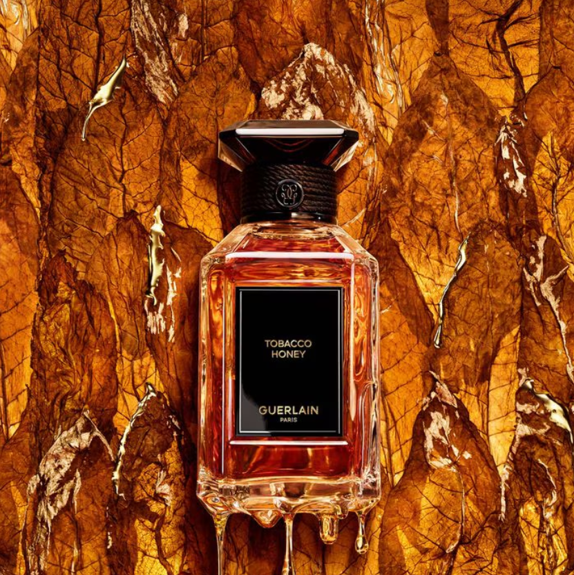 Guerlain Tobacco Honey perfume bottle centered against a textured amber and gold leaf background. The rectangular glass bottle contains amber-toned liquid and has a black front label with gold text reading 'TOBACCO HONEY' and 'GUERLAIN PARIS,' plus a black cap.
